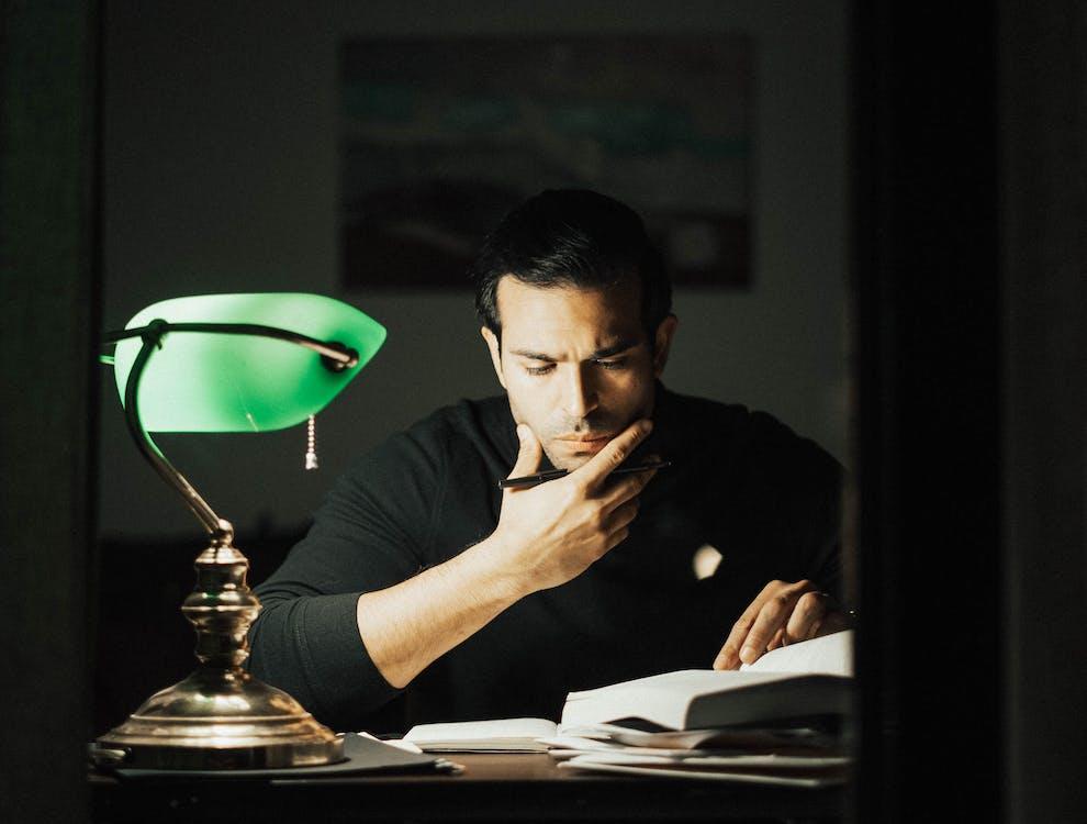 Free Serious young male wearing black turtleneck reading book and touching chin thoughtfully while studying in dark home office Stock Photo