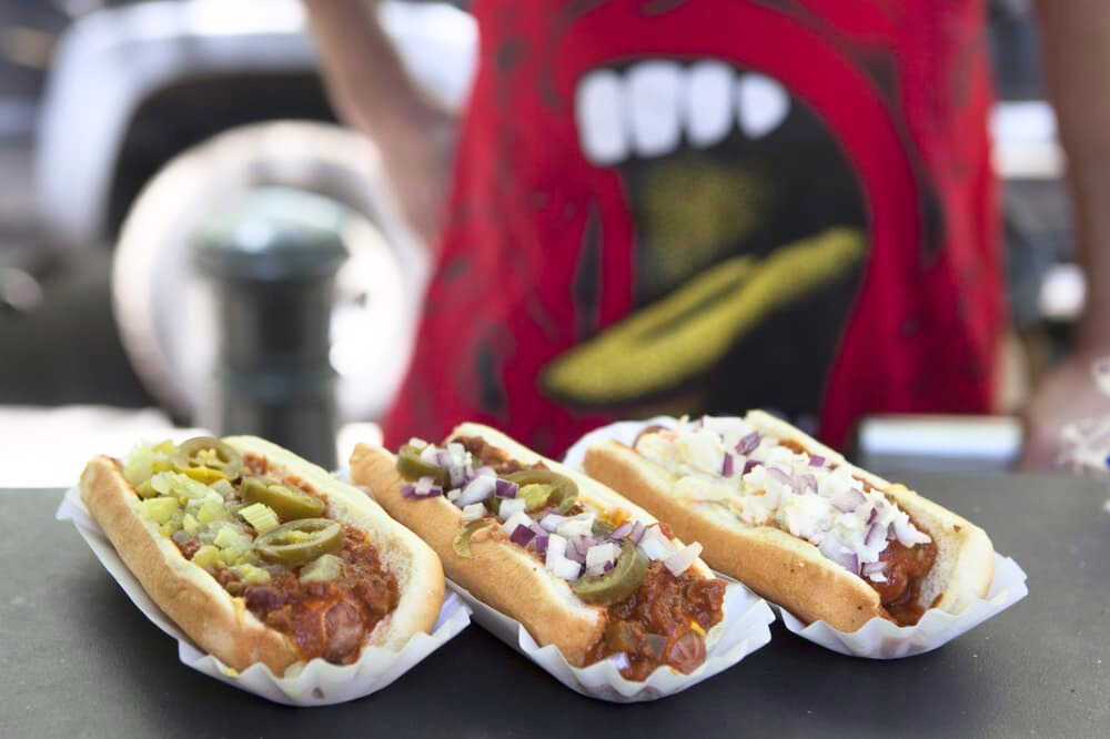 Photo from Chili Man's Yelp page. Three hot dogs, smothered in chili, jalapenos, onions, and cheese, lined up on the edge of a food cart. A man's torso wearing an apron is blurry behind.