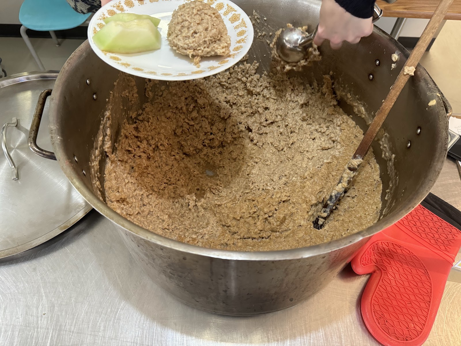 A small hand scoops from a big pot of oatmeal. There is a red oven mitt to the right.