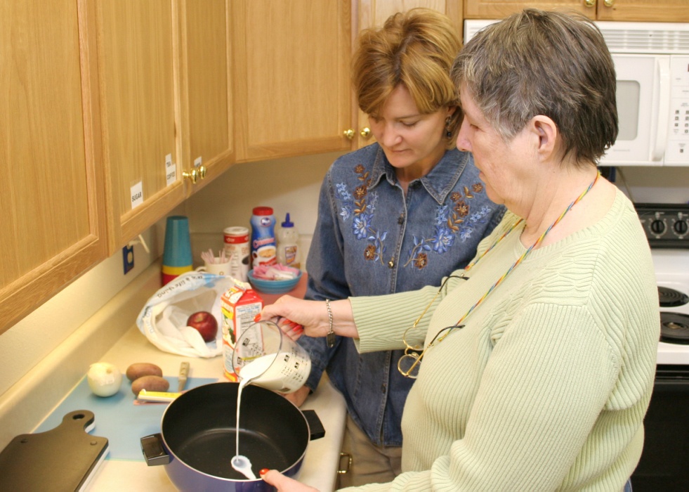 Two older women, one in a tan sweater pouring milk into a pan, one in a jean jacket helping the other woman.