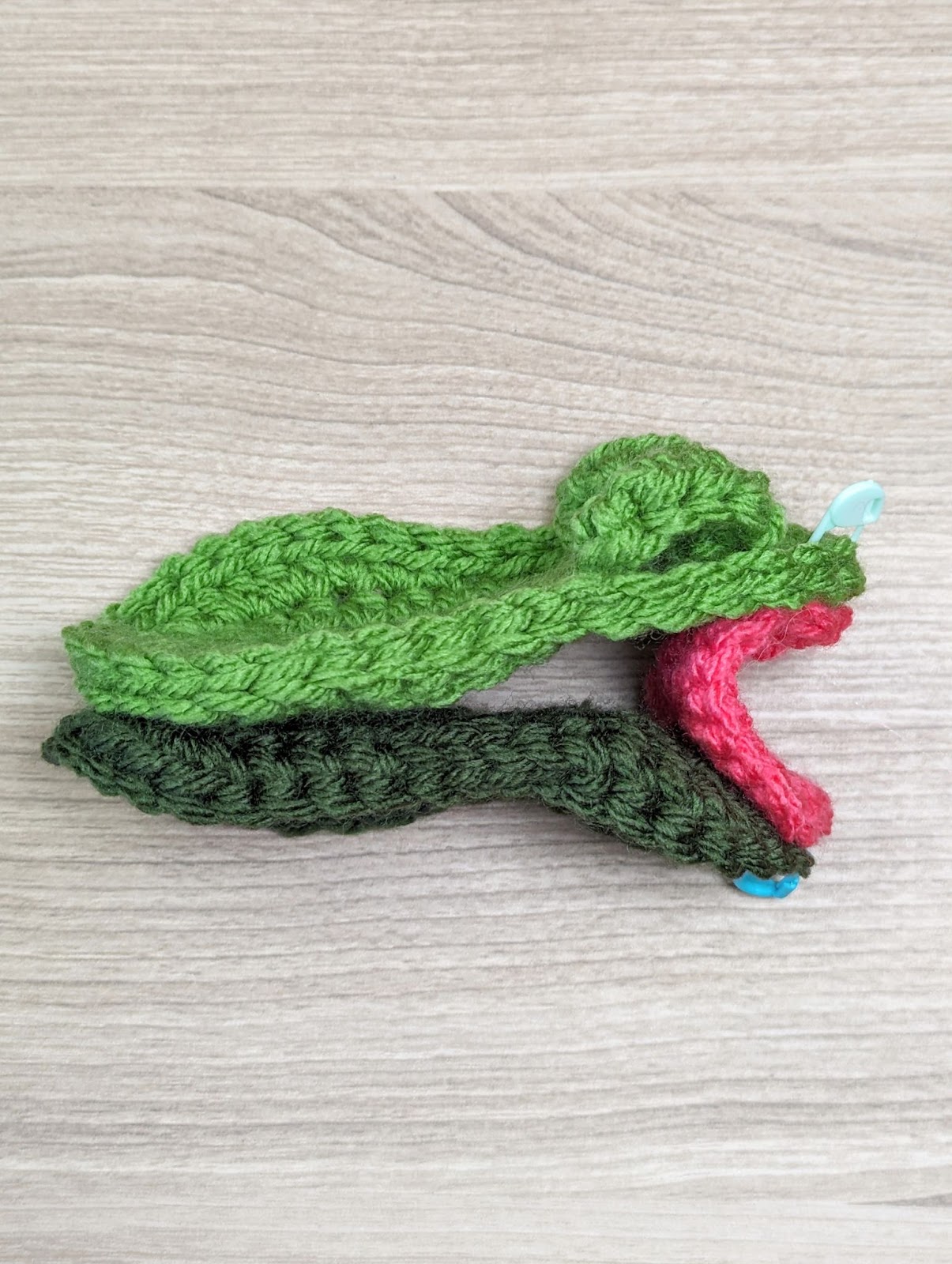 A hand-knit piece resembling the head of a frog with its mouth open, using green yarn for the head and upper jaw, darker green for the lower jaw, and pink for the mouth. The knitted object is placed on a wooden surface.