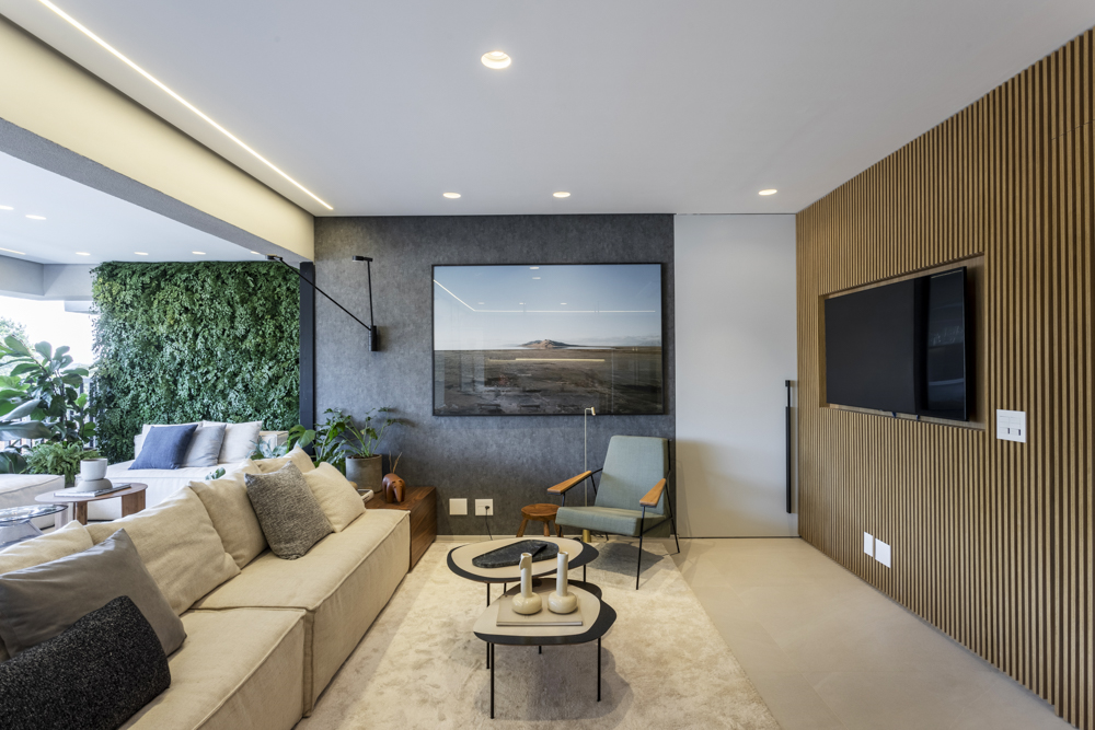 Sala de televisão com um jardim vertical que cobre parte da parede onde tem um televisor. Tem um sofá caramelo e outro cinza, duas mesinhas no centro e uma parede com detalhes em madeira.