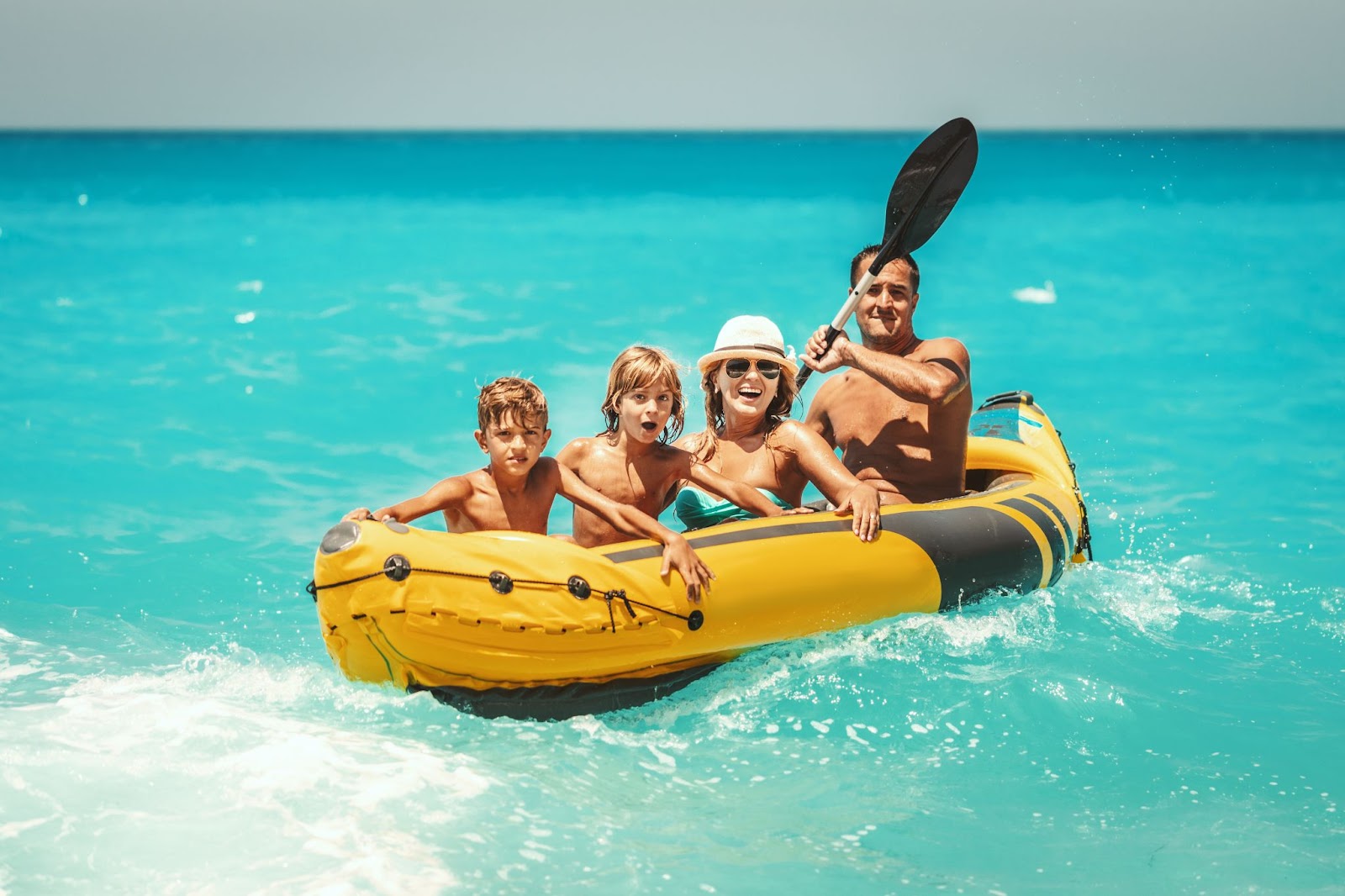 A happy family is enjoying a ride in a rubber kayak, and waves are splashing them in tropical ocean water during summer vacation.
