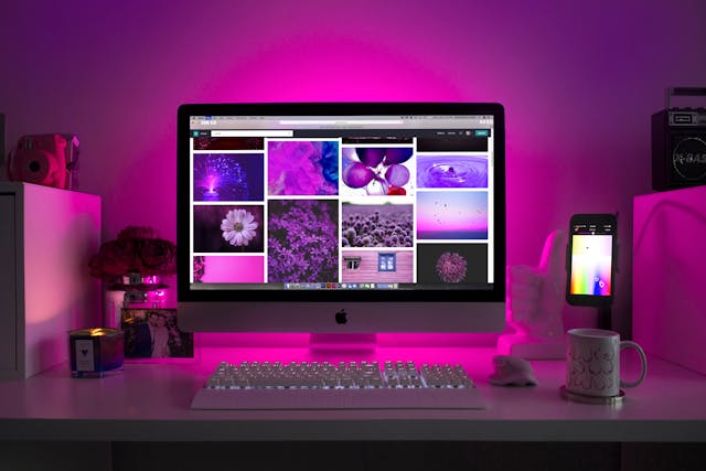 A table with a monitor keyboard and mouse with a pink light from behind the monitor illuminating the wall behind