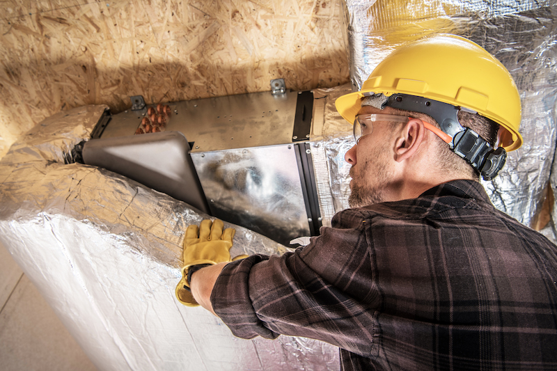 An HVAC technician in a yellow hard hat and plaid shirt inspects and repairs an air duct system in an attic, a crucial step in addressing issues when air ducts make noise due to blockages or loose fittings.