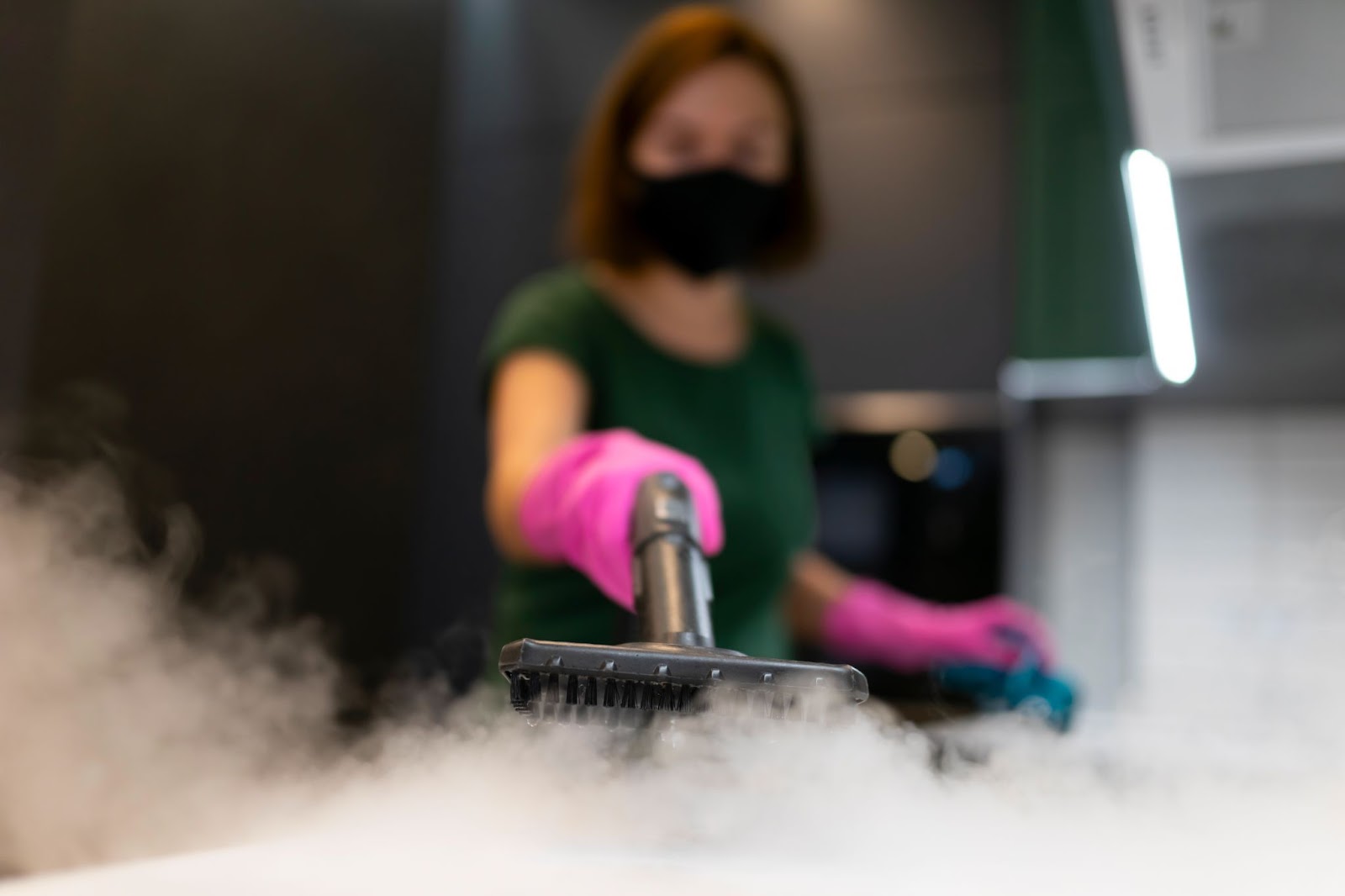 A blurred image shows a professional female cleaner in a green top and pink rubber gloves releasing vapor from a steam generator after a deep cleaning session.