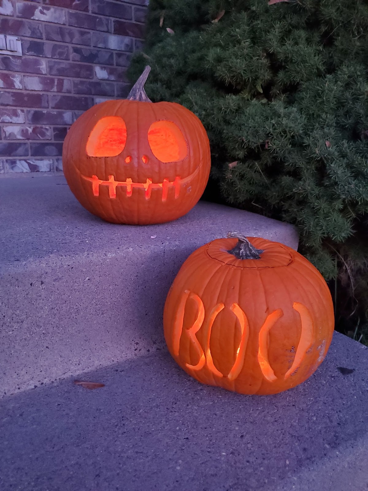 Two jack-o-lanterns on a set of concrete steps to a front porch. One is Jack Skellington and the other is carved to say “Boo”