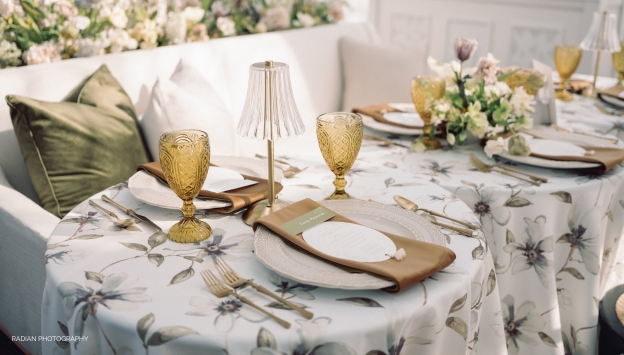 2 round set tables with white linens, yellow glasses, and green plants.