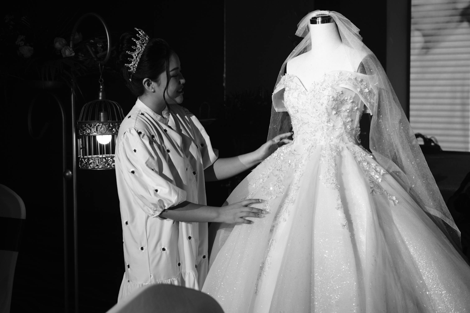 Bride in tiara adjusting elegant lace wedding dress on mannequin in black and white photography