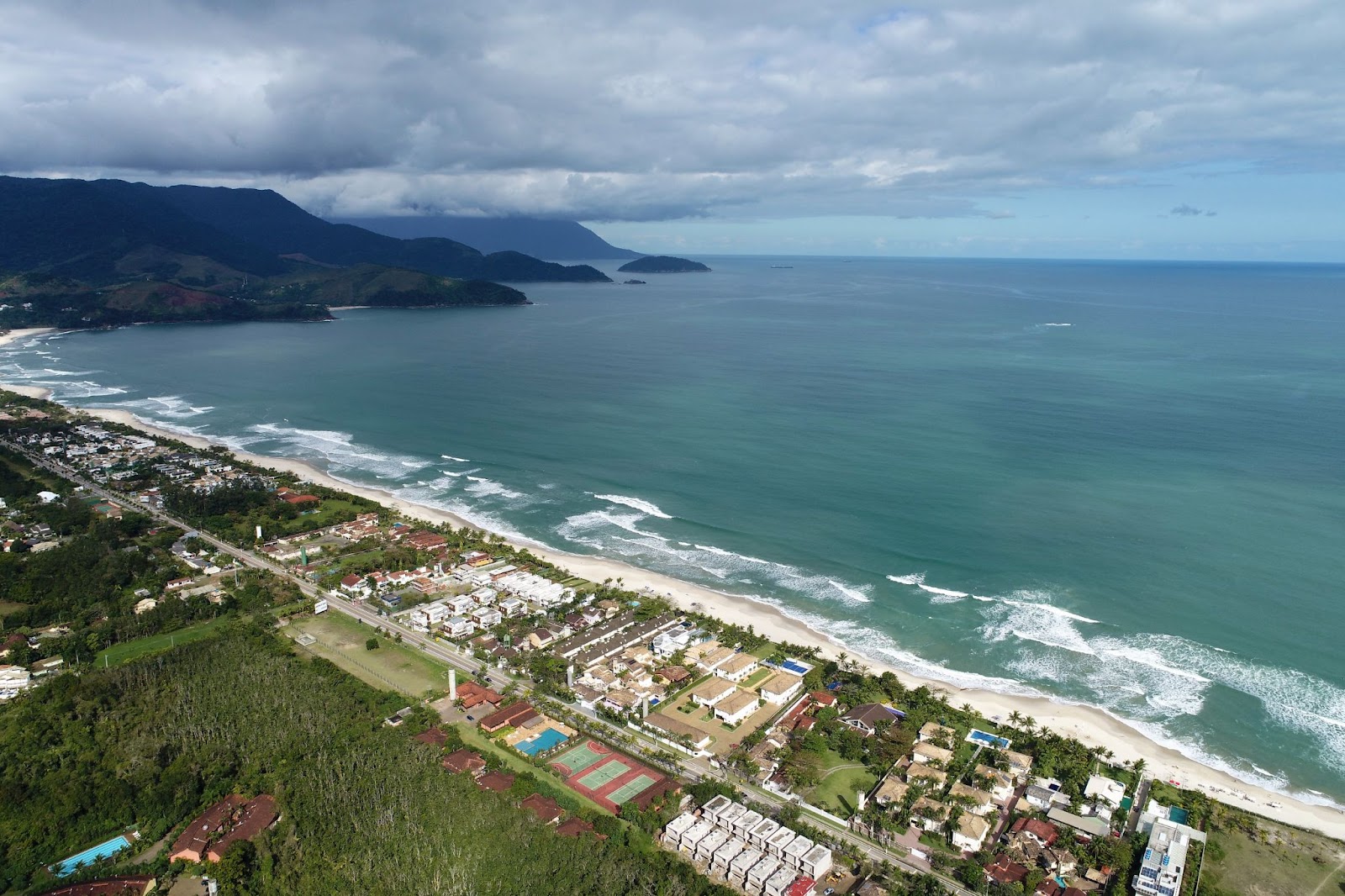 Vista aérea da Praia de Maresias, em São Sebastião. A longa faixa de areia branca é limitada pela água azul do mar e por uma fileira de construções baixas.