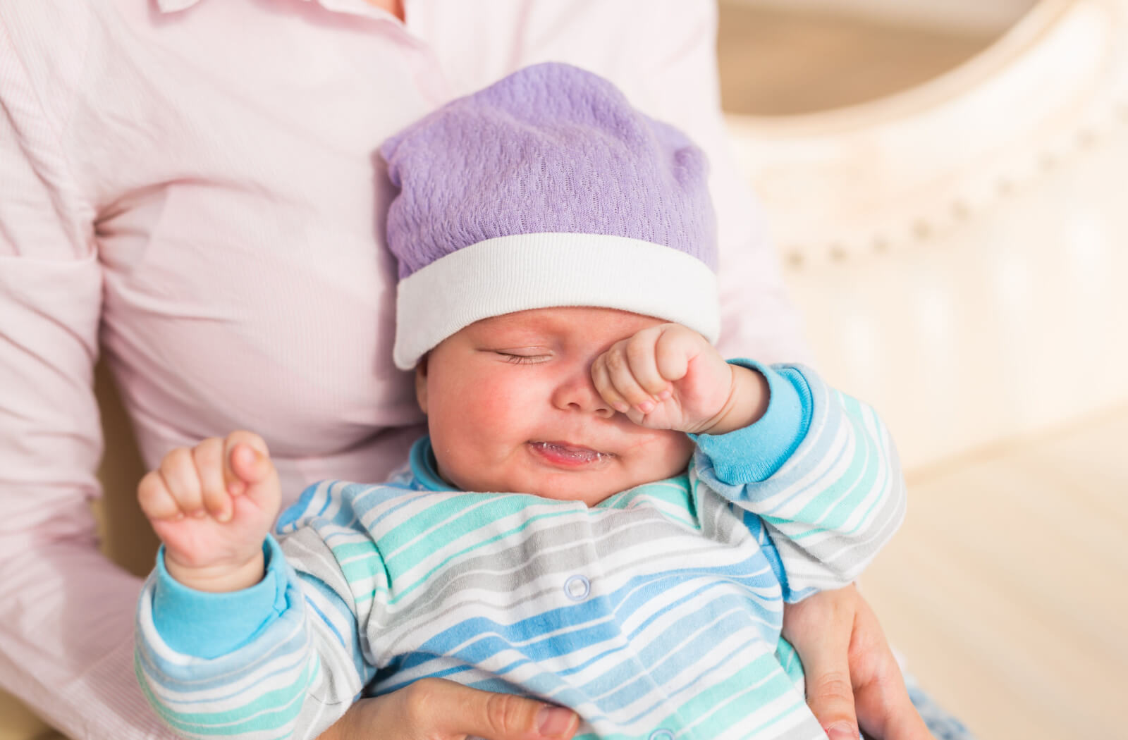 A newborn baby in striped pyjamas rubbing their eye.