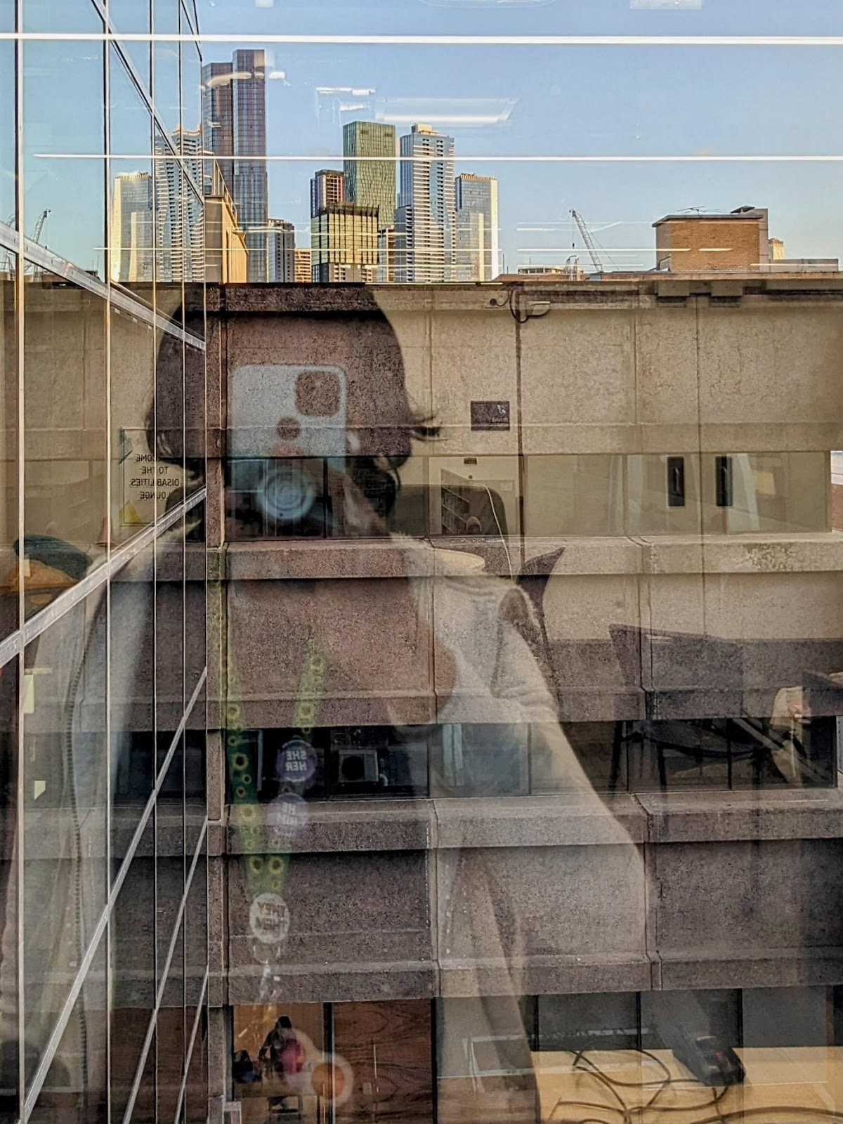 A self portrait, shot on a camera phone in the reflection of a window looking out from the Disabilities Lounge in Building 168 to the Melbourne Skyline. The phone obscures the photographer's face. They are wearing a sunflower lanyard, adorned with pins and a fidget toy that hangs amongst the keys.