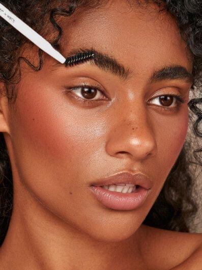 a dark skinned woman brushing her brows using a spool brush in the style of soap brows