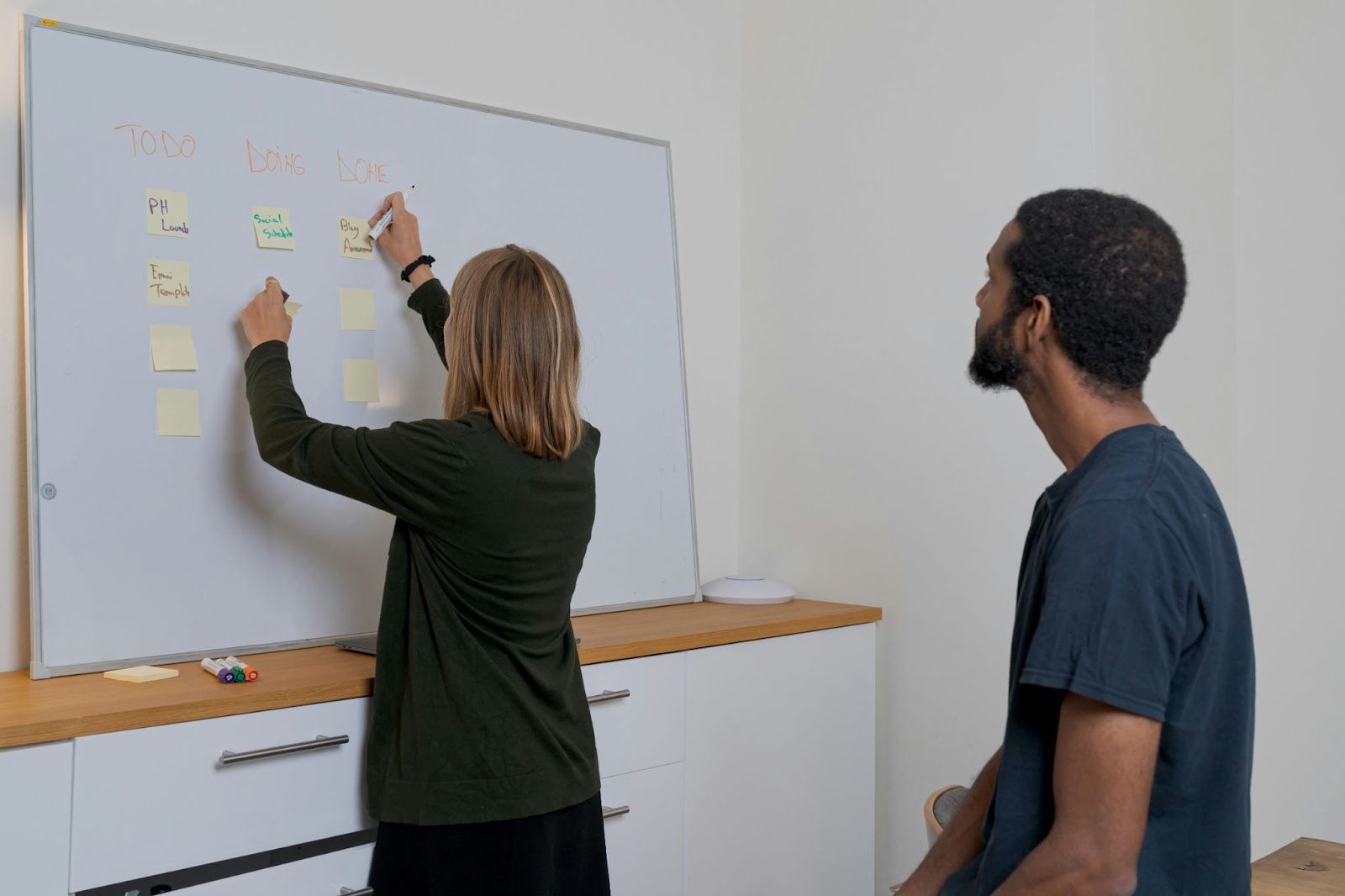 a woman placing sticky notes on a board with Todo, In progress and Done columns while a man watching attentively