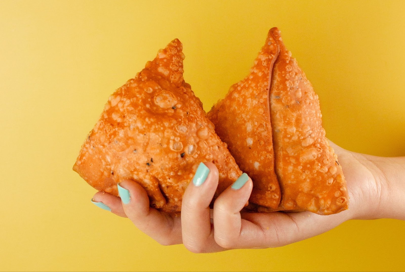 closeup of a hand with mint green nail polish, holding two samosas at a kelowna indian restaurant; against a yellow background