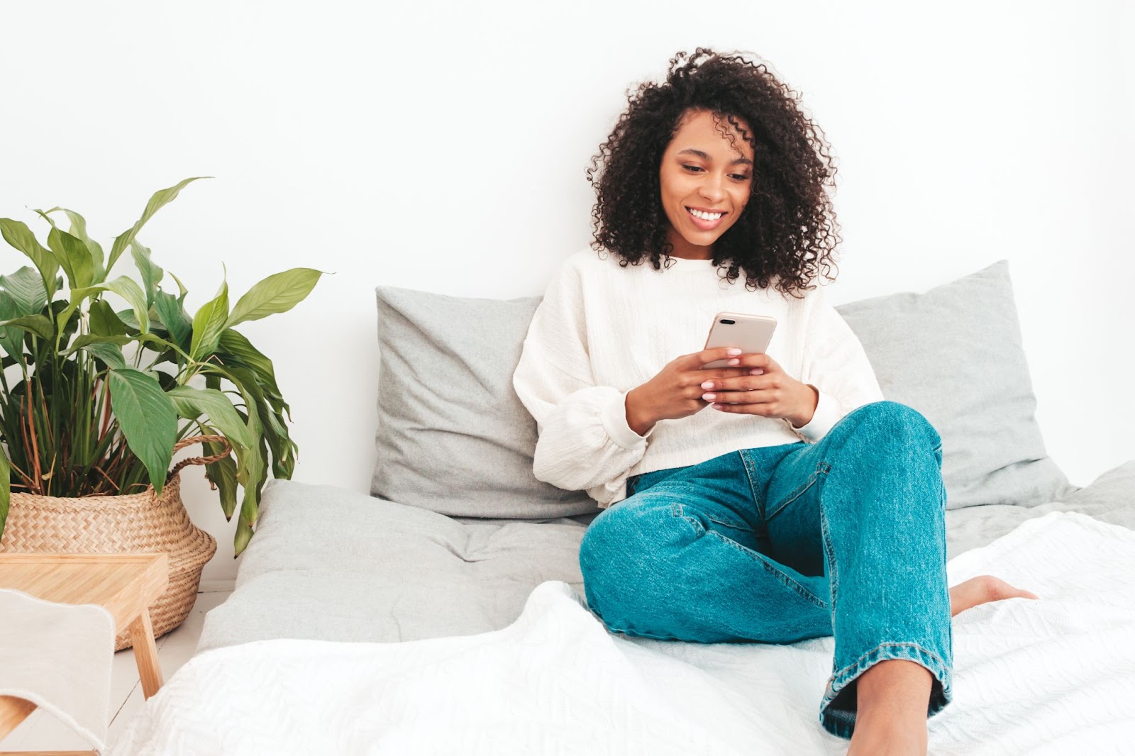 Uma mulher de blusa branca e calça jeans está sentada na cama conferindo o benefício do abono salarial em seu smartfone.