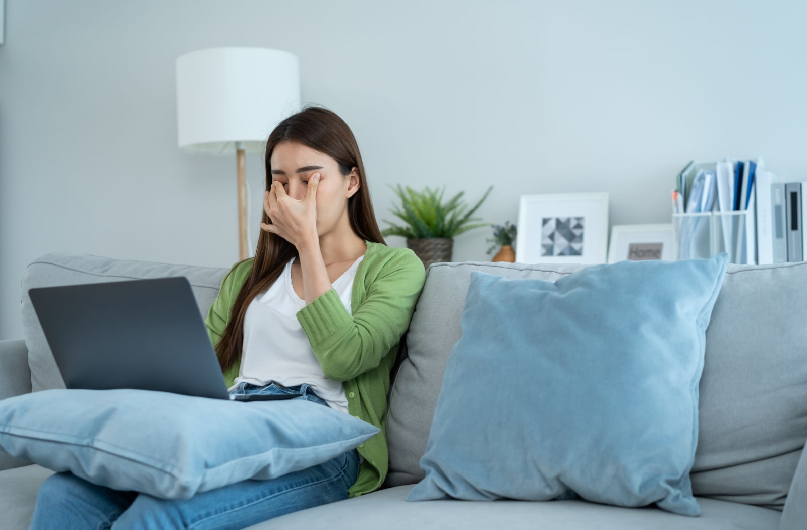 A woman sitting on a couch with a laptop sitting on her lap. She's rubbing her eyes due to eye strain