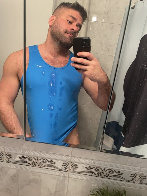 Max Romano wearing a blue tank top covered in jizz taking a photo in the mirror in the bathroom