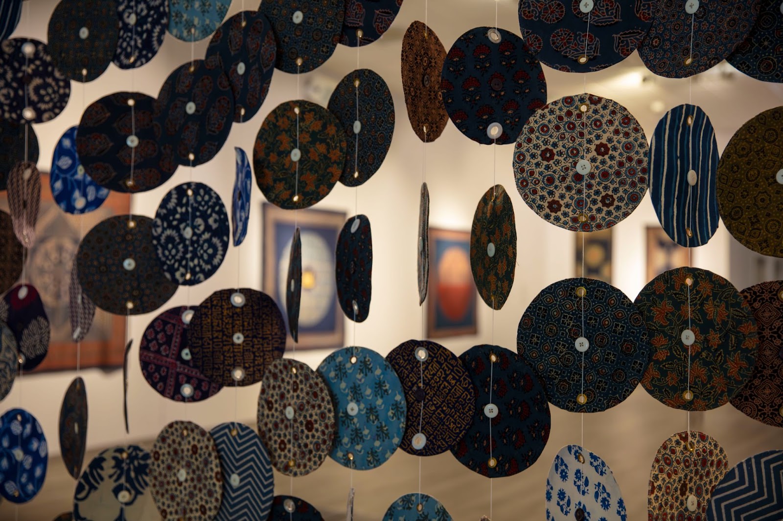 Image: Installation View, Indigo: The Blue Gold, 2023. Migrated Communities hangs on a wall of the exhibition, with Homage to the Farmers of Champaran, 1917-1918 in the foreground. The latter piece takes the shape of many patterned circles hanging in the space. Courtesy: South Asia Institute.