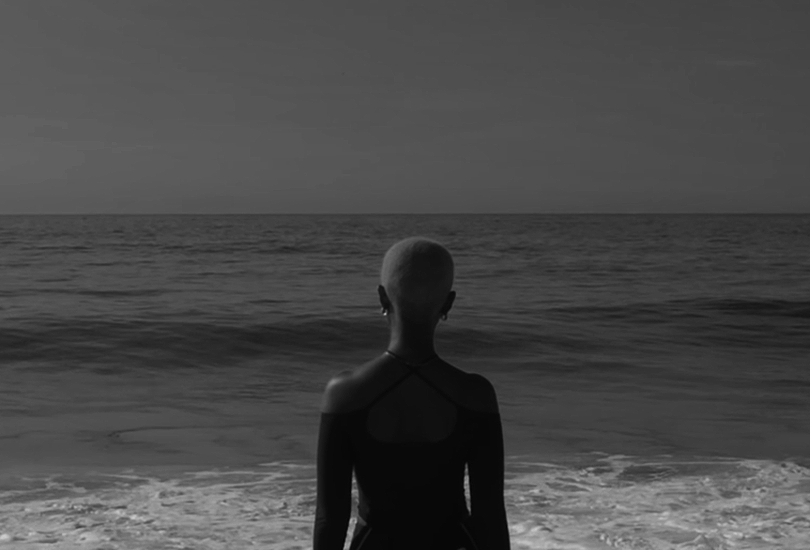 A desaturated screenshot of Kelela from her music video for "Enough for Love". Featuring a shot of her from behind, stood on the beach and staring out at the ocean.