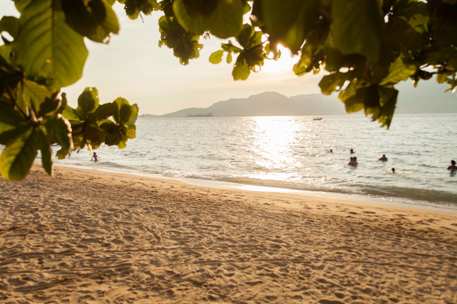 Praia do Julião, em Ilhabela, ao entardecer. A areia dourada está vazia, mas há alguns banhistas no mar. As águas estão tranquilas e refletem o brilho do sol.