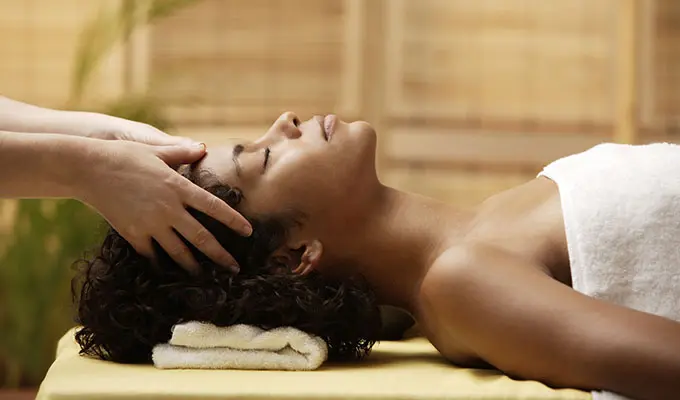 A woman in a towel is laying on a massage bed while receiving a scalp massage