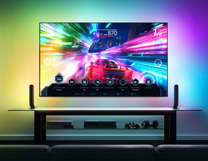 Wall-mounted Samsung TV surrounded by RGB lighting, two speakers, and a game console, displaying a racing game with the Game Bar