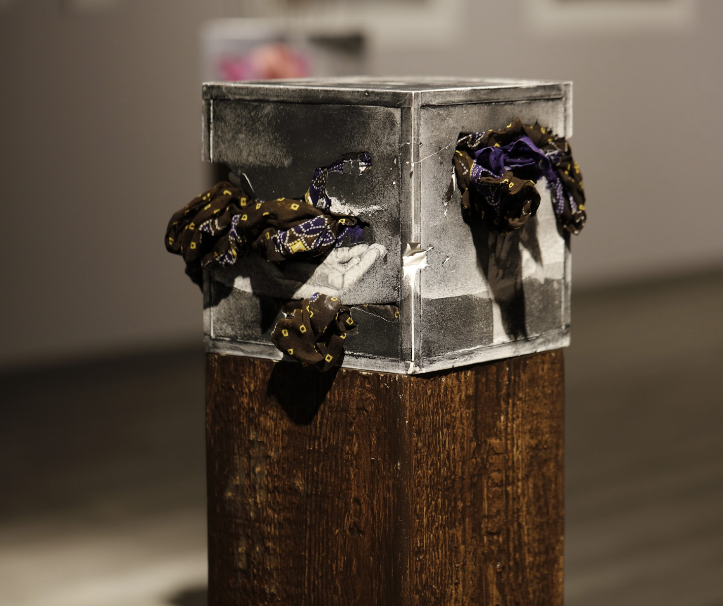 Image: Nirmal Raja, Contained III, 2017. Collagraphs on cast white plaster, sari fabric. A metallic colored cube upon a wooden plinth, the corners of the plinth and the cube flush. On two sides of the cube, black and purple fabric bursts out. Image courtesy of the South Asia Institute.