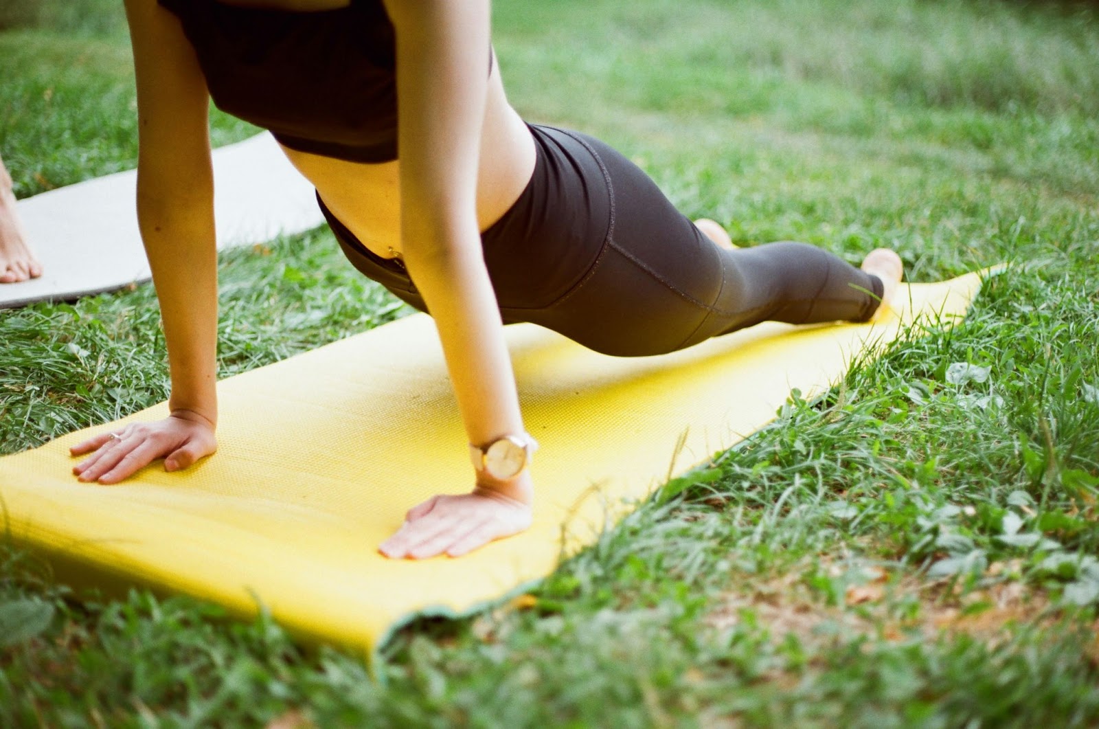 A person in black tights and sports bra doing outdoor yoga with a yellow mat on green grass.