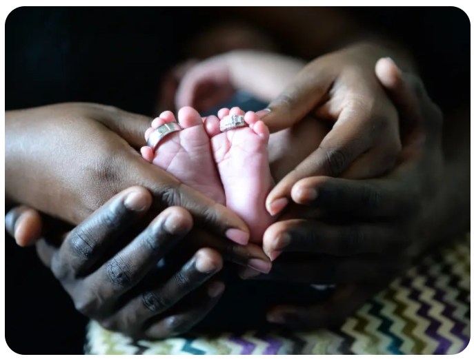 A portrait of two pairs of African American hands cradling the baby feet of an African American baby