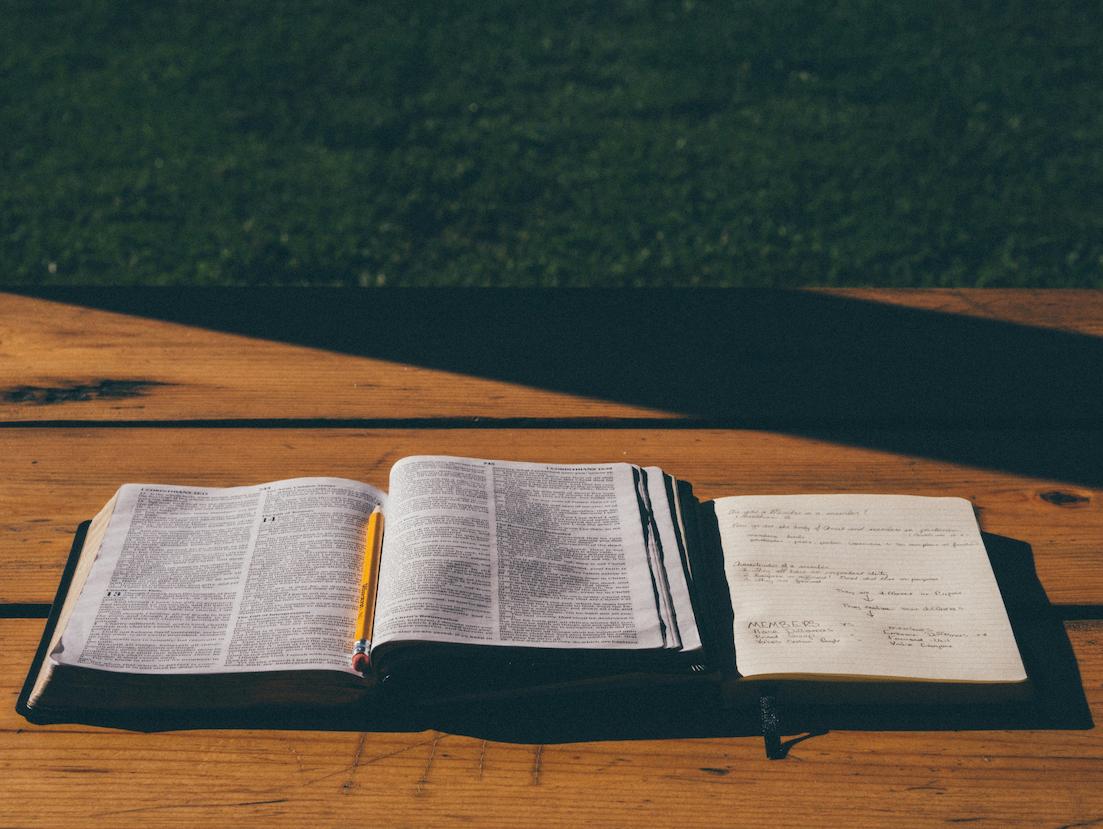 An opened Bible with a pencil on top of it and a notebook on the side with notes.