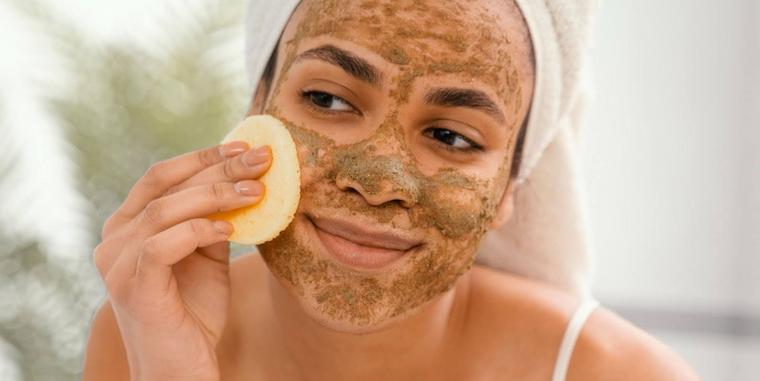 Uma mulher está fazendo spa em casa ao aplicar um esfoliante em seu rosto enquanto sorri e usa uma toalha na cabeça.