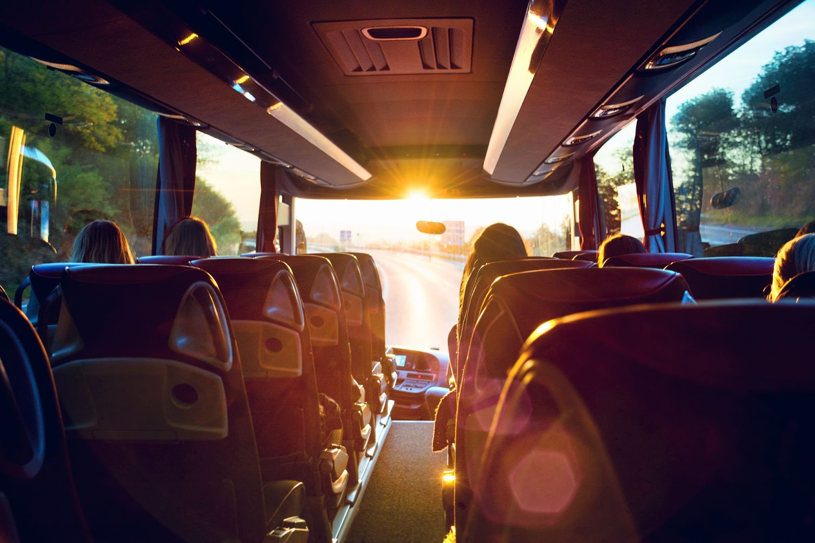 Foto de dentro de um ônibus de viagem com bancos de costas, virados de frente à estrada, com a luz do nascer do sol iluminando seu interior.