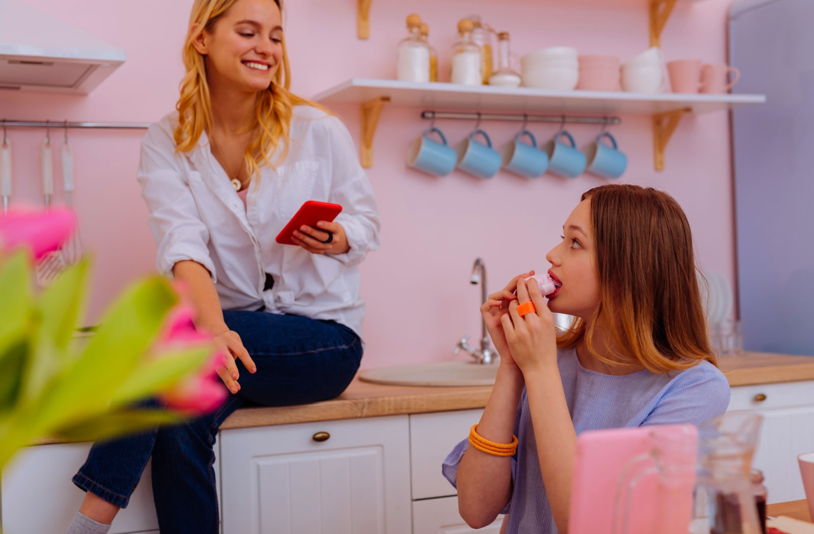 An older adult sister sitting on the counter in a kitchen and talking to her younger, teenage sister who is eating breakfast