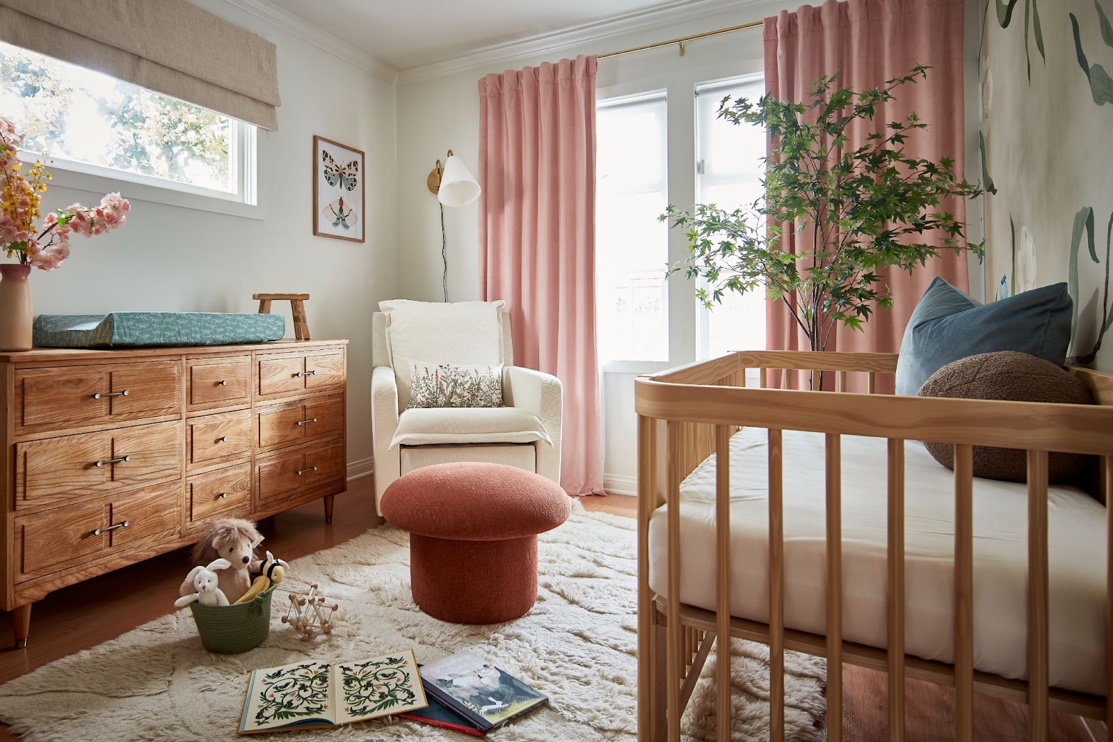 Charming kid's bedroom with a pastel pink theme. Pink curtains, ottoman, and flowers complement neutral tones. Natural wooden chest of drawers, and a warm, furry white carpet create a cozy and delightful atmosphere.