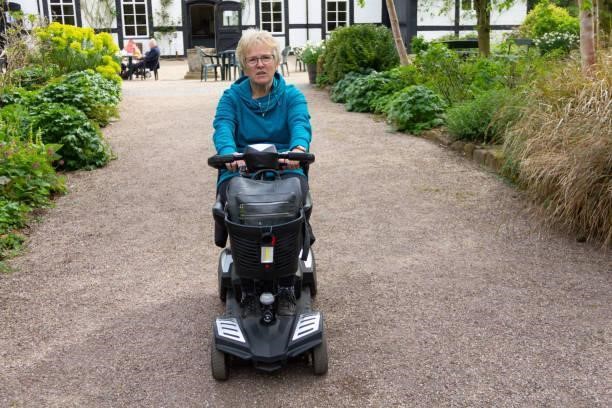 Blond woman rides her disability scooter along path between gardens enjoying the freedom and independence the scooter gives her to enjoy looking round the gardens on her own at her own pace. Blond woman rides her disability scooter along path between gardens enjoying the freedom and independence the scooter gives her to enjoy looking round the gardens on her own at her own pace. elderly scooter stock pictures, royalty-free photos & images