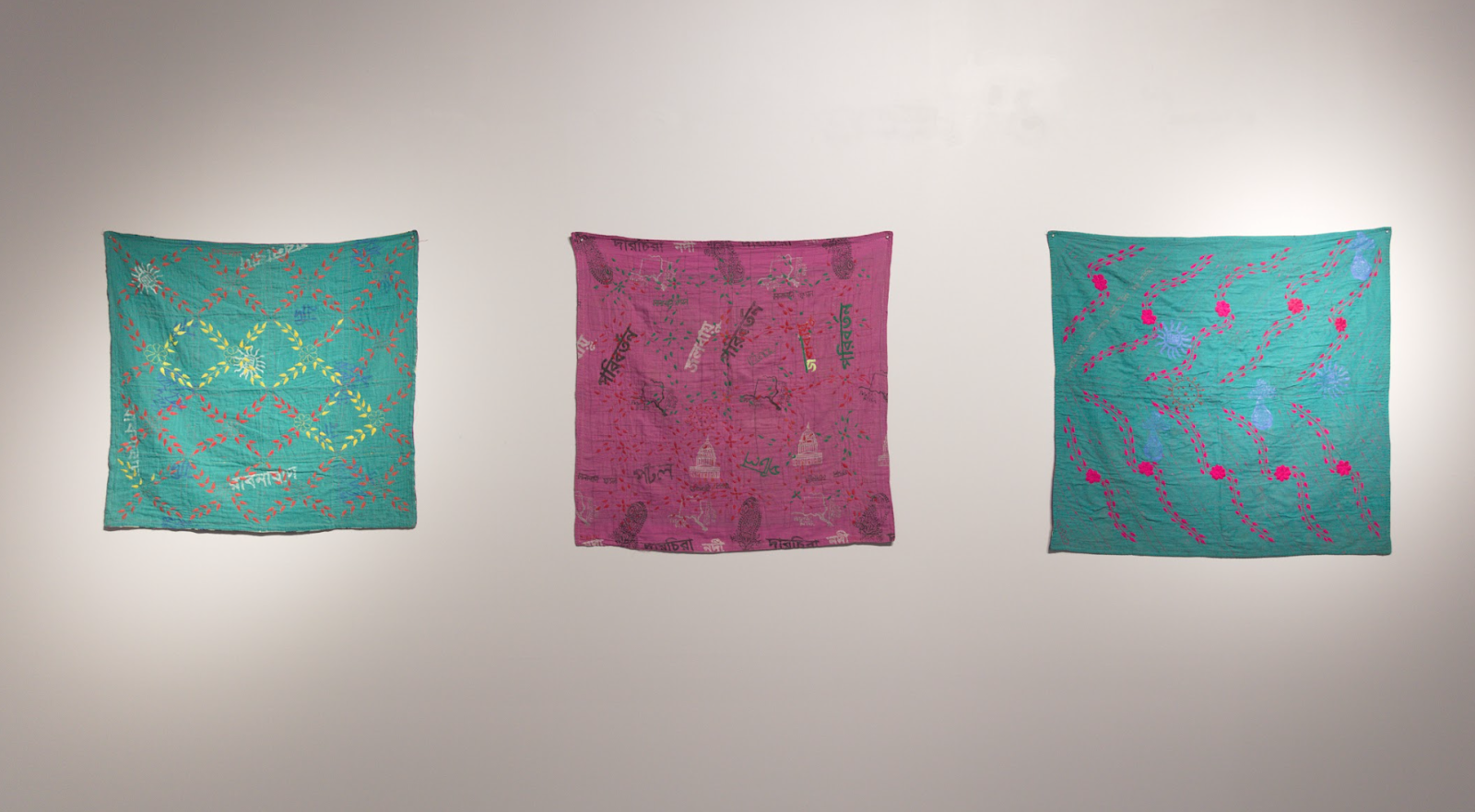 Image: Installation view, from left to right: Verdant Kantha (2021), Capitol Kantha (2021), and Hot Pink Flowers on Green Fields Kantha (2021). The three square cloths—from left to right, teal, magenta, teal—hang in a row against a white wall, with printed and embroidered patterns of different colors on them. Image courtesy of the South Asia Institute.