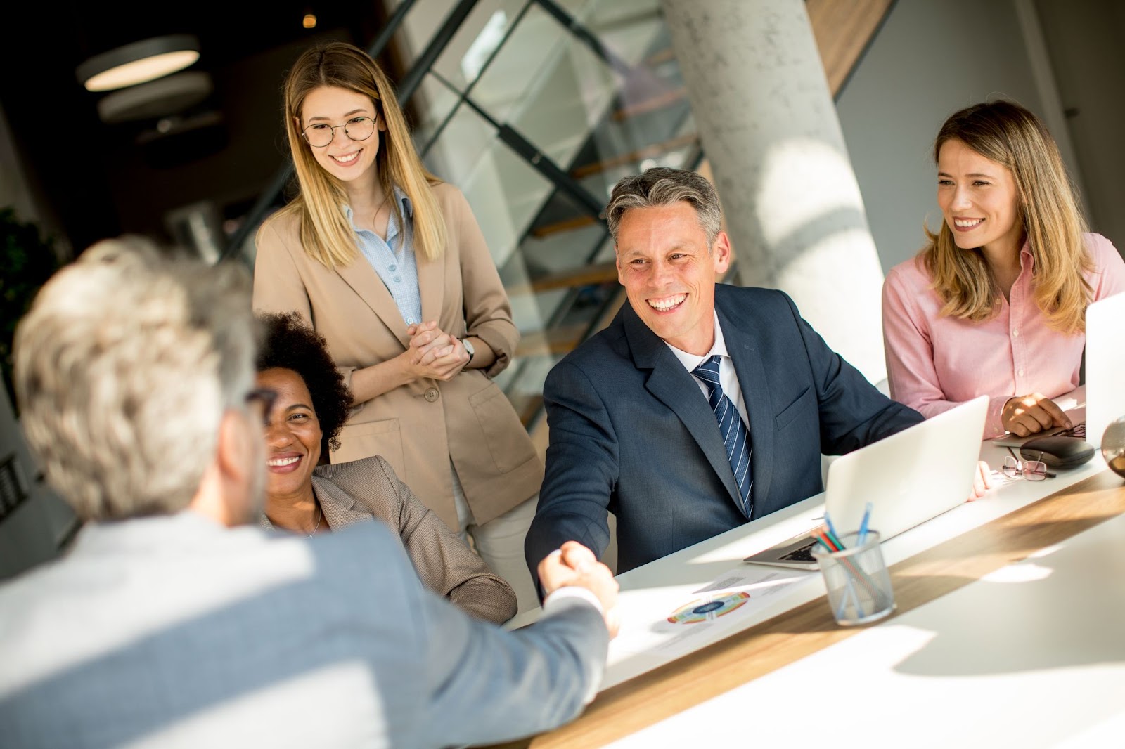 A male businessman and four women shake hands by a desk, signifying a partnership agreement with a management consulting firm.