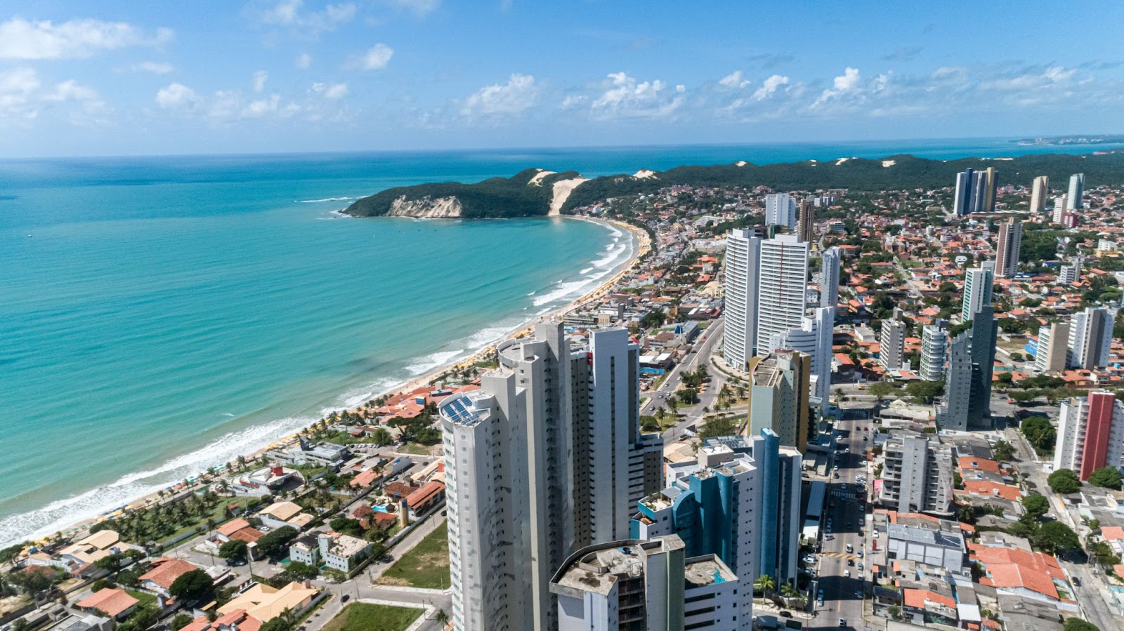 Vista aérea da cidade de Natal. Mar azul cristalino ao lado esquerdo da foto, com casas e prédios da cidade ao lado direito.