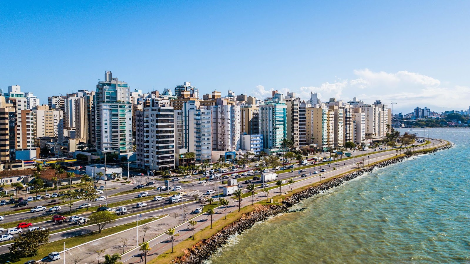 Vista aérea da Avenida Beira-Mar, em Florianópolis. A longa via onde circulam vários automóveis é limitada pelo mar e por prédios modernos de diversos tamanhos. Ao fundo, o céu aparece azul e quase sem nuvens.