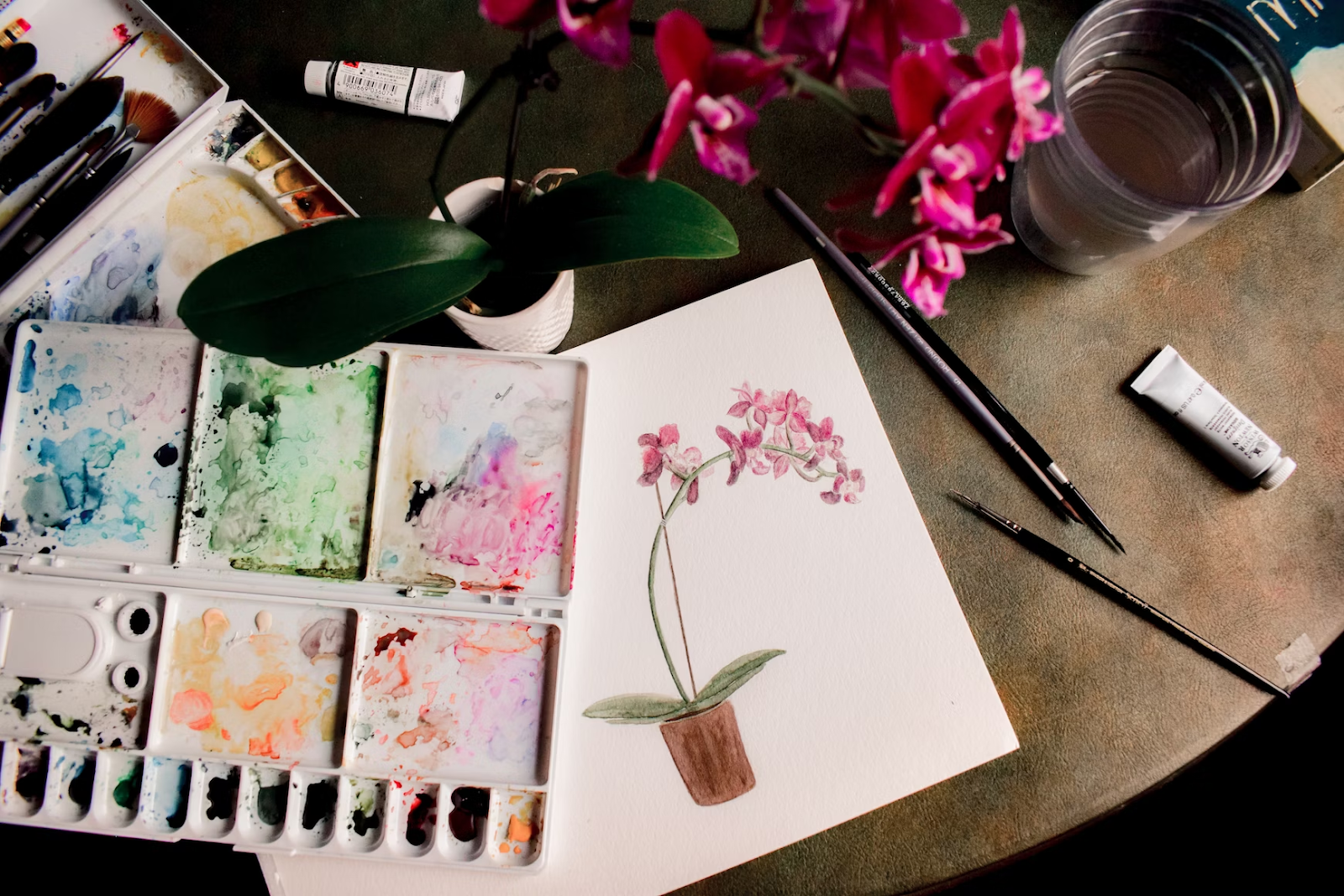 an overhead shot of an artist's brown desk with a painting of a flower pot, watercolors, brushes, cups, and flowers