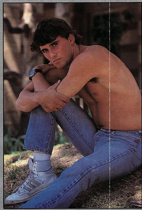 Tex Anthony posing shirtless in denim jeans and running shoes outside under a tree