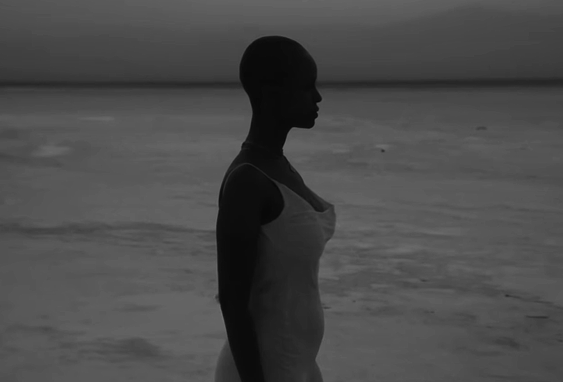 A desaturated screenshot of Kelela from her music video for "Washed Away". Featuring a shot of her stood on the shore of a beach, looking out, wearing a white dress.