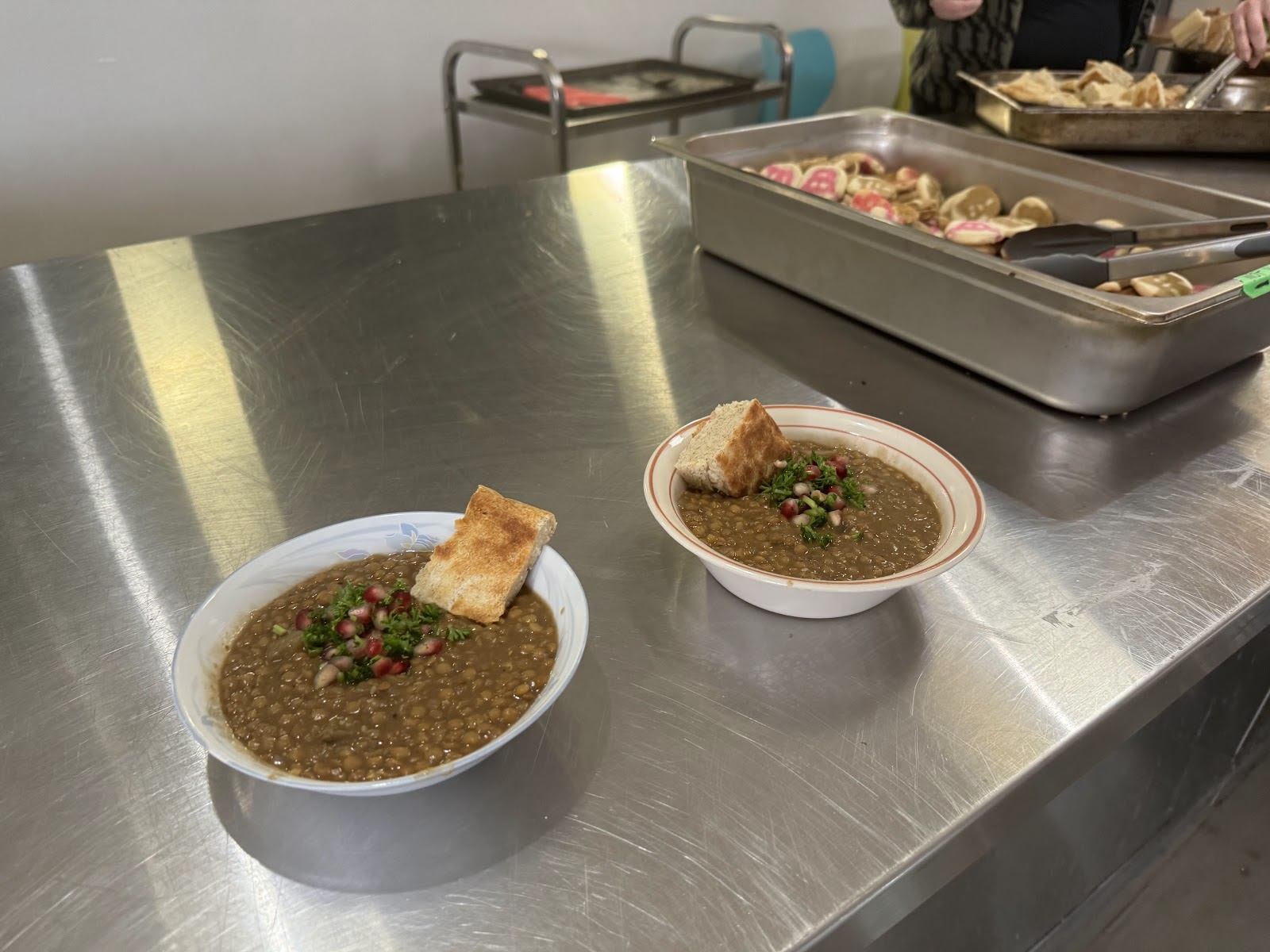 Two bowls of lentil eggplant stew with some parsley and pomegranate seeds on top, served with a piece of bread. In the background, there is a big metal container of cookies and bread.