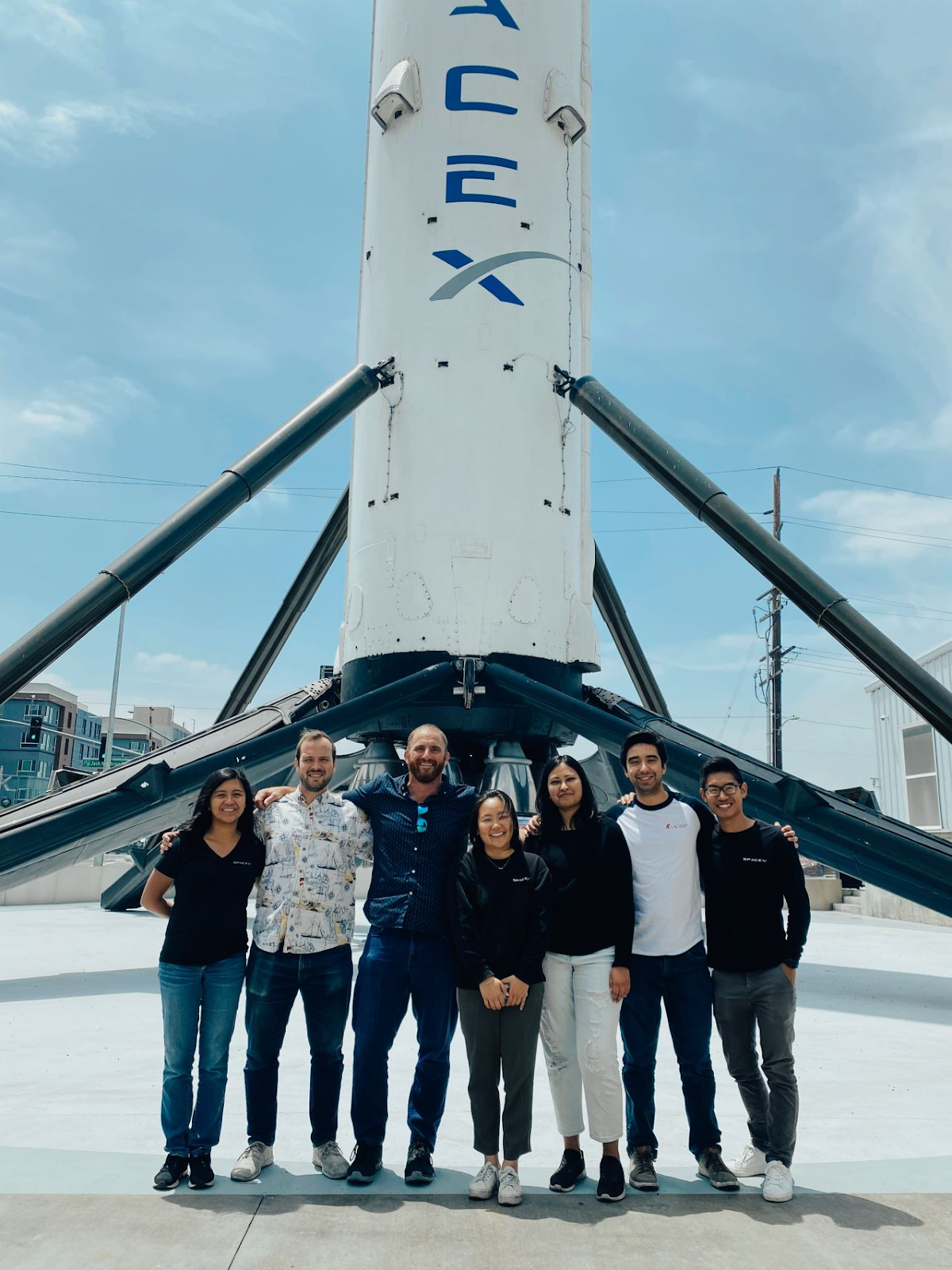 A group of coworkers pose in front of a SpaceX rocket on a sunny day.