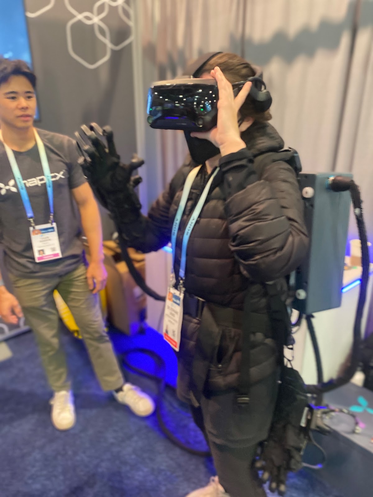 A brunette woman dressed in all black is holding black VR goggles to her face, has a haptic glove on her right hand, and is wearing on her back on a grey box with black cords