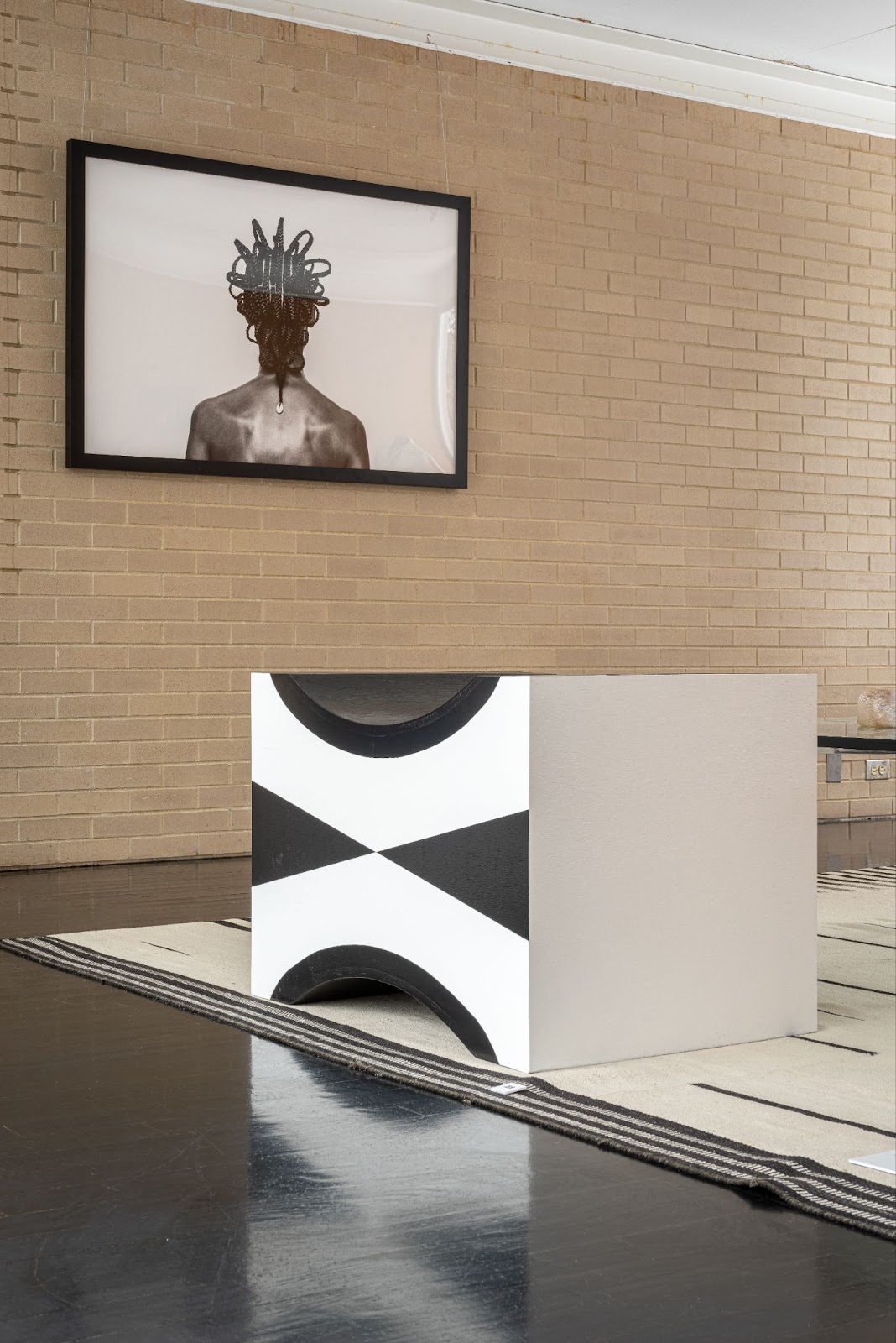 Image: Installation view of A Love Supreme: McCormick House Reimagined. A black and white cube sits on the floor of the gallery. A photograph by Shani Crowe of the back of a dark-skinned model with intricately braided hair hangs on the wall. Photo by Siegfried Mueller, courtesy of the McCormick House. 