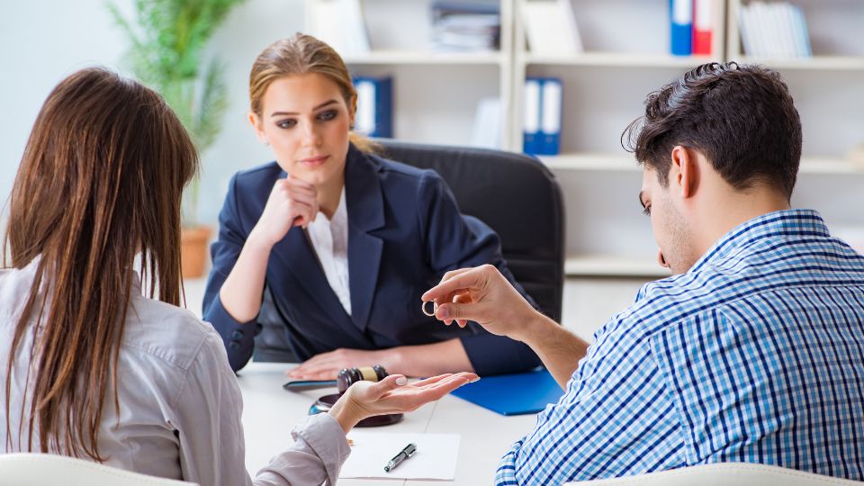 A professional setting with three individuals engaged in a meeting: a woman in a business suit sitting across the table looking attentively at a man and a woman. The man, wearing a checkered shirt, is handing a ring to the woman sitting next to him, possibly in the context of a legal or formal proceeding, as the woman in the suit appears to be overseeing the exchange.