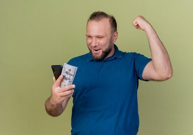 Foto grátis homem eslavo adulto alegre segurando um dólar e um celular, olhando para eles e fazendo um gesto de sim