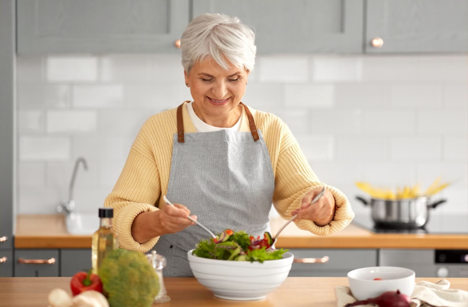 An older adult woman in a kitchen making a bowl of salad with both hands and smiling